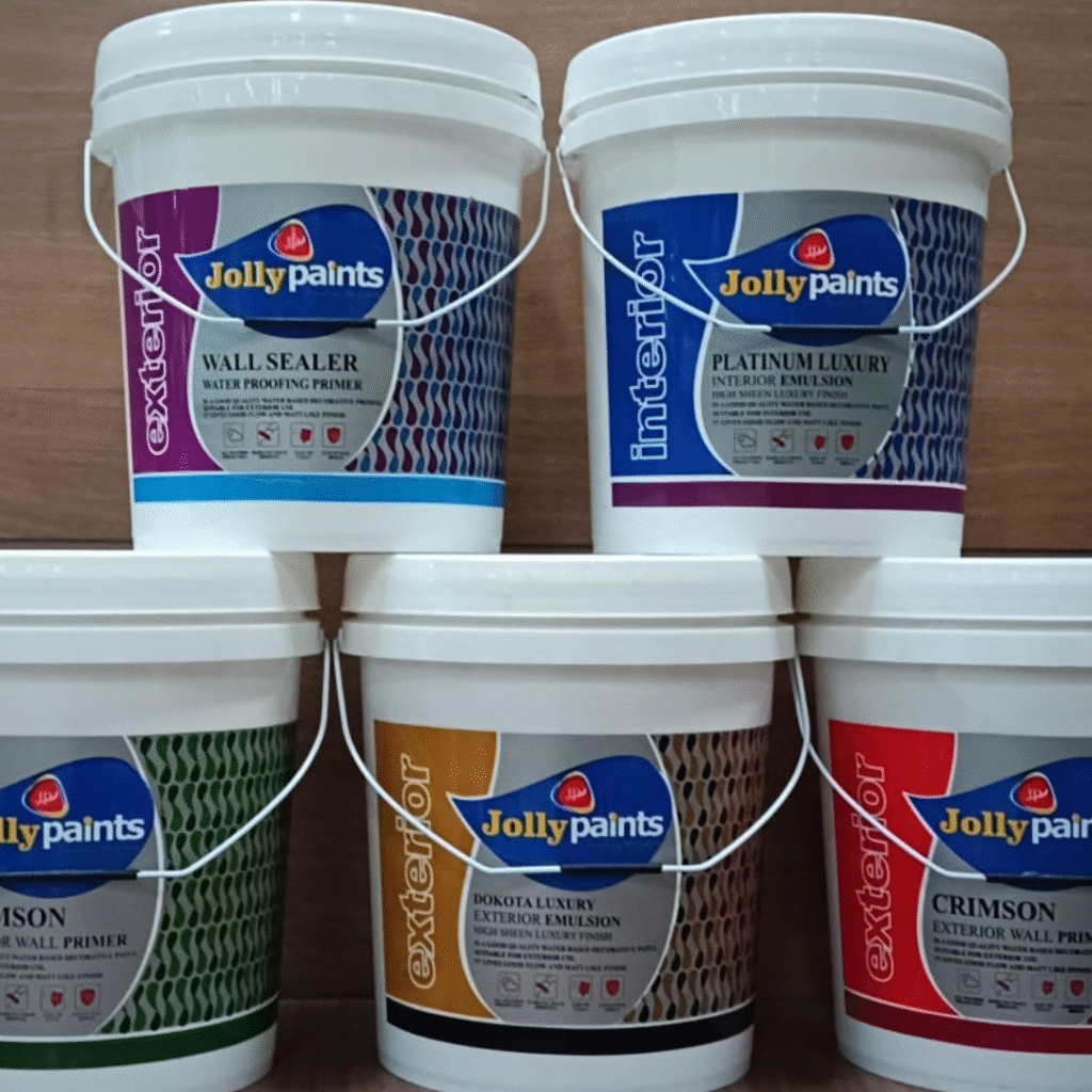 Five Jolly Paints plastic buckets arranged in a tiered layout against a wooden background. The top row features Wall Sealer Waterproofing Primer and Platinum Luxury Interior Emulsion. The bottom row displays Crimson Interior Wall Primer, Dakota Luxury Exterior Emulsion, and Crimson Exterior Wall Primer. Each bucket has bold, colorful labels with the Jolly Paints logo prominently visible."