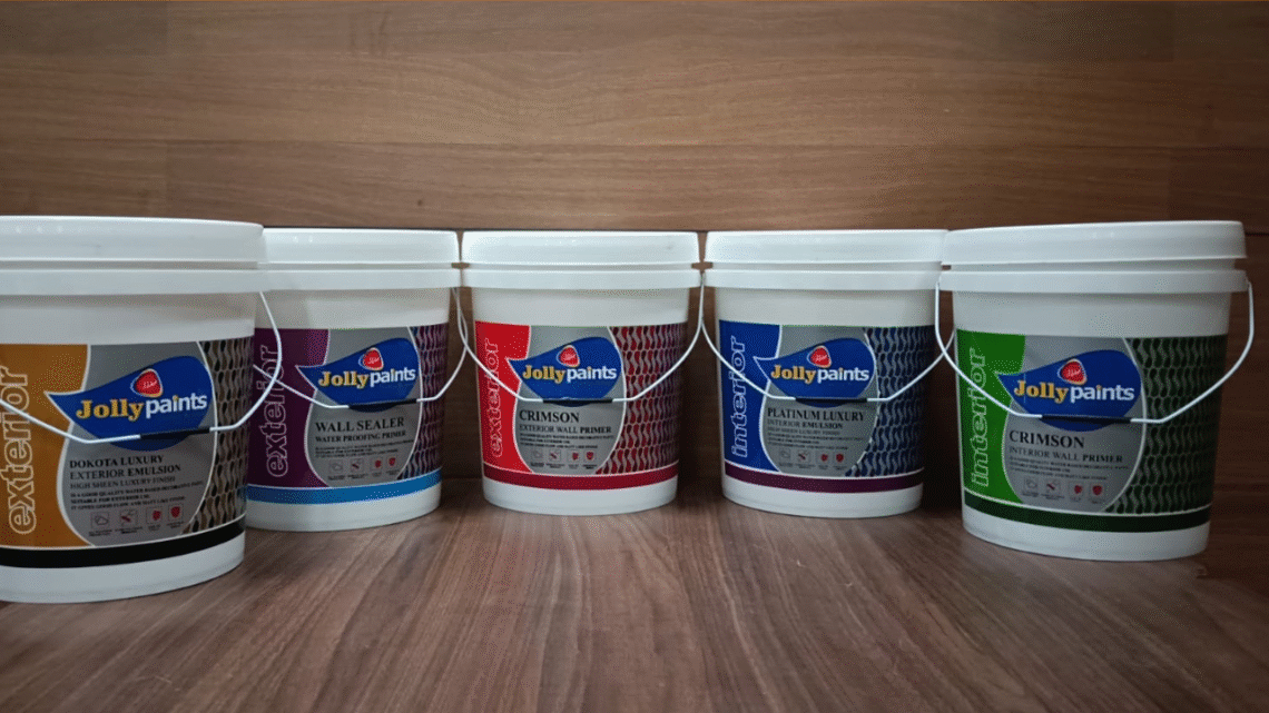 Five large white plastic paint buckets from the brand Jolly Paints are arranged in a row on a wooden surface. Each bucket has a different colored label indicating various interior and exterior paint and primer products, including Dakota Luxury Exterior Emulsion, Wall Sealer Waterproofing Primer, Crimson Exterior Wall Primer, Platinum Luxury Interior Emulsion, and Crimson Interior Wall Primer."