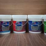 Five large white plastic paint buckets from the brand Jolly Paints are arranged in a row on a wooden surface. Each bucket has a different colored label indicating various interior and exterior paint and primer products, including Dakota Luxury Exterior Emulsion, Wall Sealer Waterproofing Primer, Crimson Exterior Wall Primer, Platinum Luxury Interior Emulsion, and Crimson Interior Wall Primer."