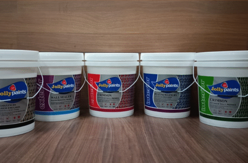 Five large white plastic paint buckets from the brand Jolly Paints are arranged in a row on a wooden surface. Each bucket has a different colored label indicating various interior and exterior paint and primer products, including Dakota Luxury Exterior Emulsion, Wall Sealer Waterproofing Primer, Crimson Exterior Wall Primer, Platinum Luxury Interior Emulsion, and Crimson Interior Wall Primer."