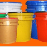 Multiple large plastic buckets in various colors—green, brown, yellow, red, blue, and white—are arranged together against a bright orange background. Each bucket has a matching or contrasting lid and industrial-style design."